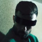 A man with futuristic eyewear poses against a textured wall, casting shadows.