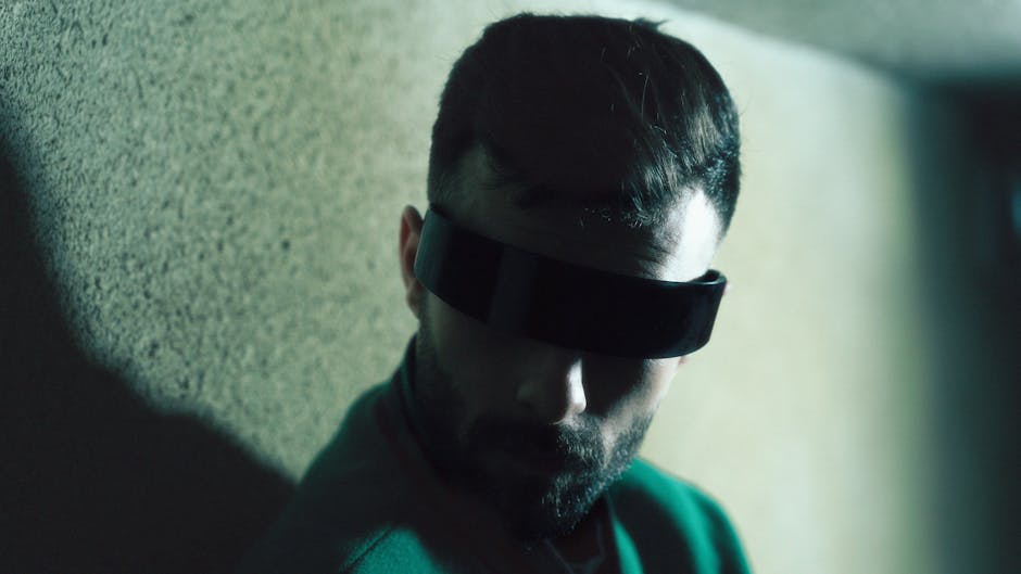 A man with futuristic eyewear poses against a textured wall, casting shadows.