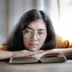 Exhausted ethnic female student in eyeglasses lying on table with stacks of textbooks while working on assignment and looking at camera
