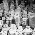 A monochrome display of various grains and spices with price tags at a market stall.