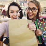 Two women smiling with shopping bags in a mall environment.