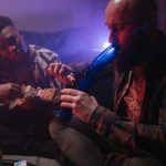 Two friends enjoying music and smoking indoors, capturing a relaxed atmosphere with a musical instrument and smoke.