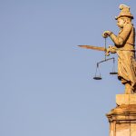 A side view of a justice statue with a sword and weighing scales against blue skies.