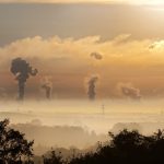 Smoke rising from factory chimneys at sunrise, symbolizing pollution and environmental impact.