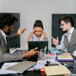 Two men argue while a woman looks frustrated at a laptop in an office environment.