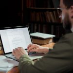 A man focuses on typing a document in a warm, cozy library environment, surrounded by books.