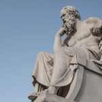 Stone statue of philosopher Socrates sitting in thoughtful pose in Athens, Greece under a clear blue sky.