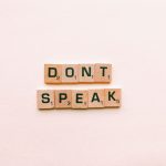 Wooden Scrabble tiles spelling 'Don't Speak' on a pink background, emphasizing silence.