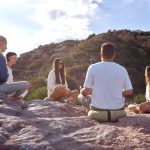 A group of adults meditating peacefully outdoors on rocky terrain under a clear sky.