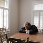 An adult man with emotions of sadness and anxiety sitting alone at a wooden table near windows.