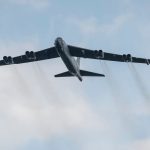 Large military aircraft flying through clear sky, emitting smoke trails from engines.
