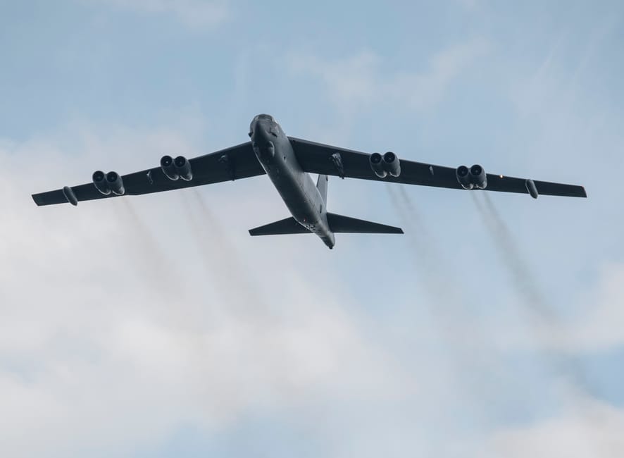 Large military aircraft flying through clear sky, emitting smoke trails from engines.