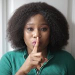 Portrait of a woman with afro hair signaling silence by holding a finger to her lips.