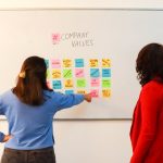 Two women discussing company values on a whiteboard in an office setting, emphasizing teamwork.