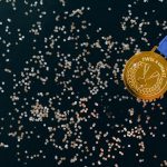Close-up of a gold medal with blue ribbon and confetti on a black surface, symbolizing victory.