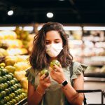 Woman wearing mask selecting fresh produce in a supermarket aisle.